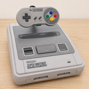 Puede incluir: Una consola Super Nintendo Entertainment System gris con un mando a juego encima. La consola cuenta con un interruptor de encendido, botón de expulsión, botón de reinicio y dos puertos para mandos. El mando tiene una cruceta, select, start y cuatro botones de acción de colores etiquetados como X, Y, A y B.