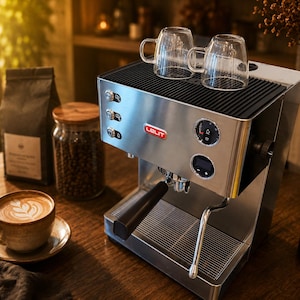 May include: A stainless steel Lelit espresso machine with a black portafilter handle, a steam wand, and a digital display. Two clear glass cups sit on the top warming tray. The machine features three circular buttons on the left and a pressure gauge.