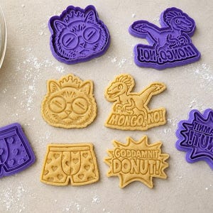 May include: A set of purple plastic cookie cutters and their corresponding dough shapes. The designs include a cat wearing glasses and a crown, a dinosaur with the text MONGO, NO!, and boxer shorts with hearts, plus a burst shape reading GODDAMNIT DONUT!