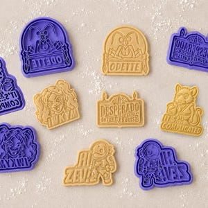 May include: A collection of purple and tan plastic cookie cutters featuring various designs including a character named Odette, a rat with the text IT'S COMPLICATED, a sign reading DESPERADO CLUB SO FUN IT HURTS, a person with IWKYM, and an astronaut with HI ZEV.