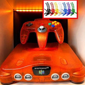 May include: A translucent orange Nintendo 64 game console and matching controller with a black logo plate. An inset image shows eight plastic controller stands in black, red, yellow, white, blue, purple, and green colors for display purposes.