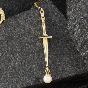 May include: Gold earring with a sword charm and a pearl dangle. The earring is hanging from a gray rock.