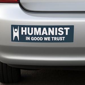 May include: A rectangular bumper sticker with the word "HUMANIST" in white capital letters above the phrase "IN GOOD WE TRUST" on a dark blue background. A white stick figure is on the left side of the sticker. The sticker is affixed to a silver car bumper.