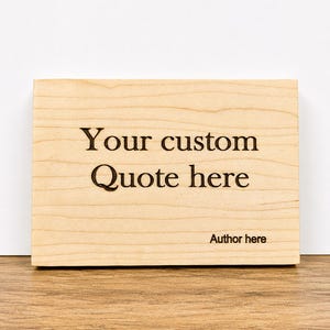 May include: A rectangular wooden sign with the text "Your custom Quote here" and "Author here" engraved in black. The sign is made of light-colored wood and rests on a wooden surface.