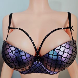 K&ouml;nnte beinhalten: Ein BH im Meerjungfrauen-Design mit Schuppenmuster in Lila- und Blaut&ouml;nen. Er hat schwarze Tr&auml;ger mit kleinen, glitzernden Details und Schleifen. Das Design umfasst einen zentralen Verschluss und eine einzigartige Tr&auml;geranordnung.