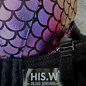 K&ouml;nnte beinhalten: Nahaufnahme eines BHs mit einem Schuppenmuster in Lila, Rosa und Gold. Der BH hat schwarze Tr&auml;ger und ein schwarzes Band. Auf dem Etikett steht "HIS.W - UK36D EURO 80DD-".