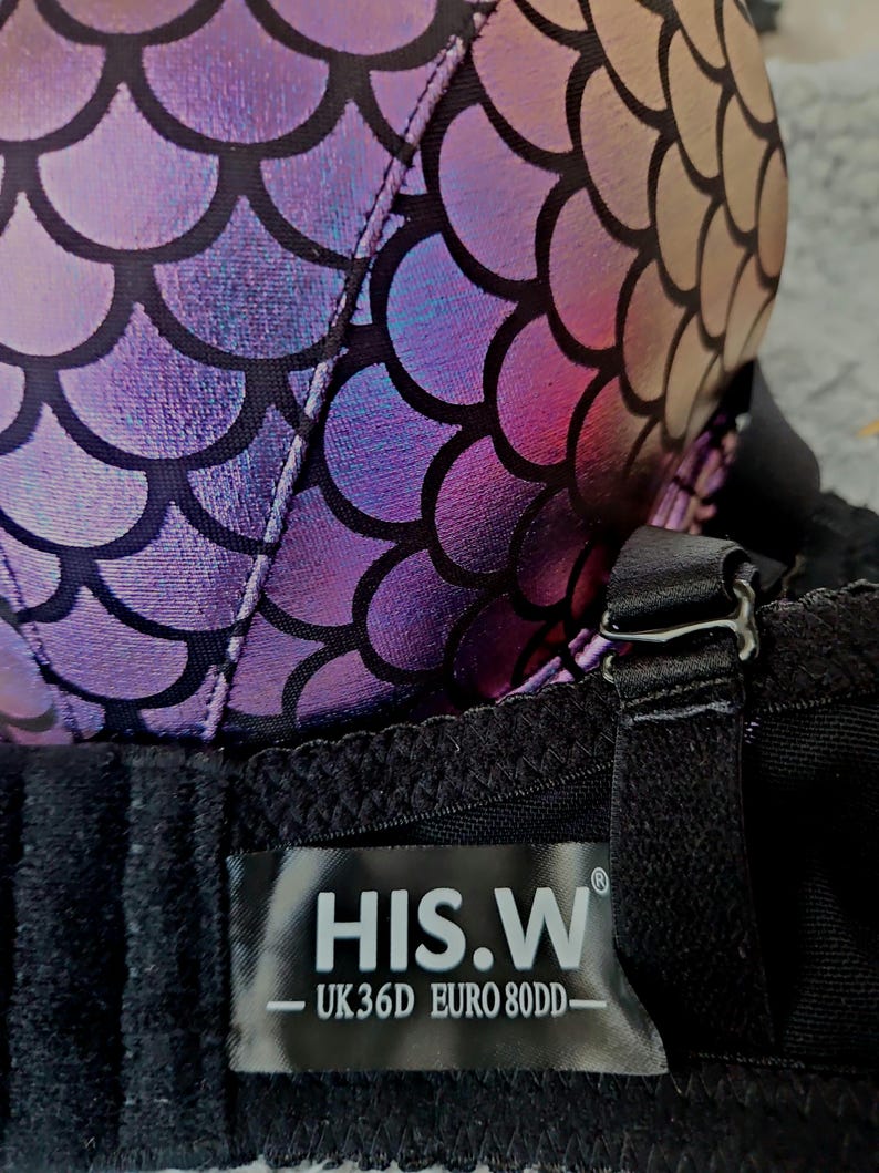 K&ouml;nnte beinhalten: Nahaufnahme eines BHs mit einem Schuppenmuster in Lila, Rosa und Gold. Der BH hat schwarze Tr&auml;ger und ein schwarzes Band. Auf dem Etikett steht "HIS.W - UK36D EURO 80DD-".