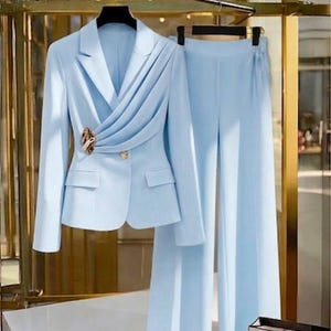 May include: A light blue women's suit set, including a blazer and matching pants. The blazer features a draped design across the front, a gold-colored brooch, and two front pockets. The pants are wide-legged. The suit is displayed on a hanger.