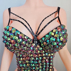 May include: A black bustier top adorned with iridescent, circular rhinestones. The top features black straps and a small square buckle. The rhinestones reflect a spectrum of colors, creating a dazzling effect. This garment is designed for special events.