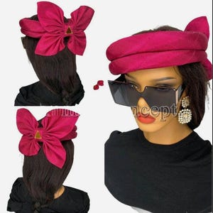 Può includere: Un turbante in tessuto magenta con un grande fiocco in cima. Il turbante è piegato a metà e ha una texture liscia. Il fiocco è fatto dello stesso tessuto e ha una forma triangolare al centro. Il turbante è indossato da una persona con i capelli scuri.