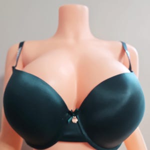 May include: A dark teal bra with a smooth, satin-like finish. The bra has underwire support, adjustable straps, and a small decorative bow with a charm in the center. The bra is displayed on a mannequin.