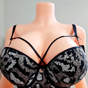May include: Black sequined bra with silver accents. The bra features a crisscross design and adjustable straps. The cups are covered in sequins, creating a shimmering effect. A small gold-colored clasp is centered between the cups.