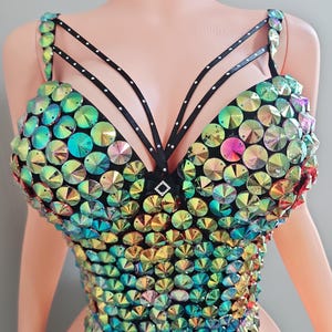 May include: A dazzling, iridescent bustier top. The top is covered in large, round, colorful rhinestones. Black straps with small white dots and a small black bow accent the design. The bustier is a vibrant piece of festival or stage wear.