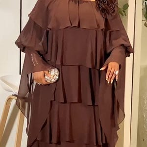 Może przedstawiać: Brązowa sukienka z odkrytymi ramionami, warstwowymi falbanami i długimi, zwiewnymi rękawami. Sukienka posiada duży materiałowy kwiat na ramieniu i jest połączona z wysadzaną klejnotami kopertówką i naszyjnikiem. Odpowiednia na formalne okazje.
