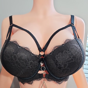 May include: Black lace bra with a decorative harness design. The bra features scalloped lace edging, adjustable straps, and a gold-colored chain detail. The bra is displayed on a mannequin.