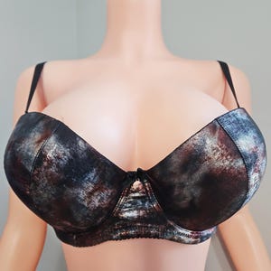 May include: A bra with a metallic, abstract pattern in shades of black, brown, and silver. The bra has adjustable black straps and a small bow detail at the center. The bra is displayed on a mannequin.