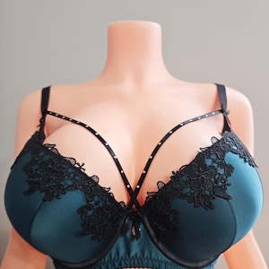 May include: Teal-colored bra with black lace detailing and a strappy design. The bra features a satin finish and is adorned with small, decorative embellishments along the straps. The lace trim adds a delicate contrast to the smooth fabric.