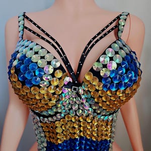 May include: A bedazzled top with a black base, adorned with iridescent sequins in shades of blue, gold, and silver. The top features thin black straps and a decorative embellishment at the center, creating a glamorous and eye-catching design.