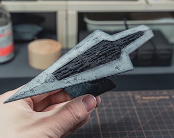 May include: A detailed model of a spaceship, painted in shades of gray and black, with a textured surface. The model is held in a hand, showcasing its intricate design and craftsmanship. The spaceship has a distinct angular shape.