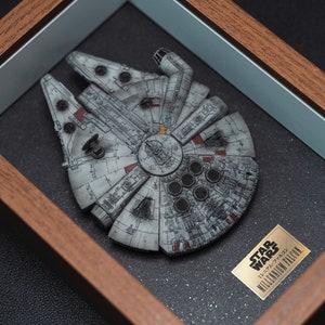 May include: A framed model of the Millennium Falcon spaceship from Star Wars. The model is painted in a weathered gray and has a black background. The frame is made of wood and has a silver mat.