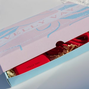 May include: A pink and blue box with the text "Velly Vay" and "We've been waiting for you to open" on the lid. The box is open and contains a red phone case with the text "Velly Vay" and a red rope.