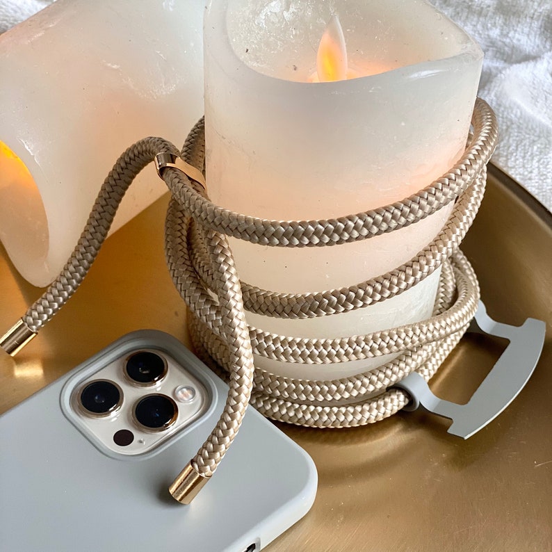 May include: A light gray phone case with a braided beige cord and gold-colored metal accents. The cord is wrapped around a white candle. The phone case has three camera lenses.