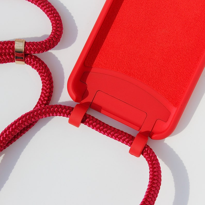 May include: A red phone case with a red braided phone strap. The strap has a gold metal ring.