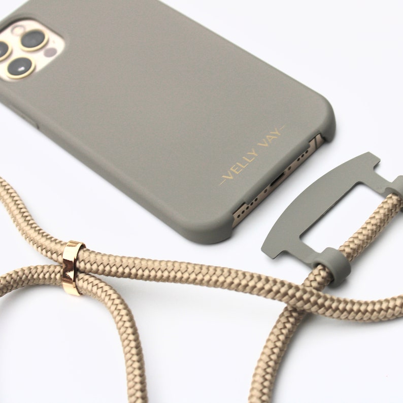 May include: A gray phone case with a matching lanyard. The case has gold text that reads "VELLY VAY". The lanyard is a beige rope with a gold-colored metal ring.