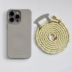 May include: A gray phone case and a coiled phone strap. The phone case has a matte finish and the text "VELLY VAY" in gold. The phone strap is a mix of pink, yellow, and gray rope with gold-colored metal accents.