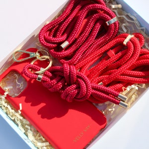 May include: A red phone case with a red rope phone strap and a gold key chain. The case has the text "Velly Vay" printed on the back.