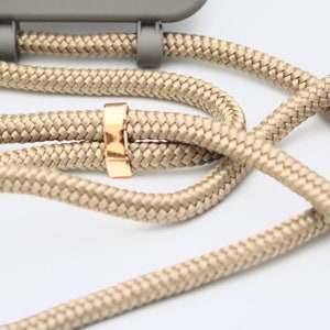 May include: Close-up of a beige woven phone strap with a gold-colored metal ring. The strap is attached to a gray phone case. The woven cord is thick and durable, and the gold ring adds a touch of elegance. The background is white.