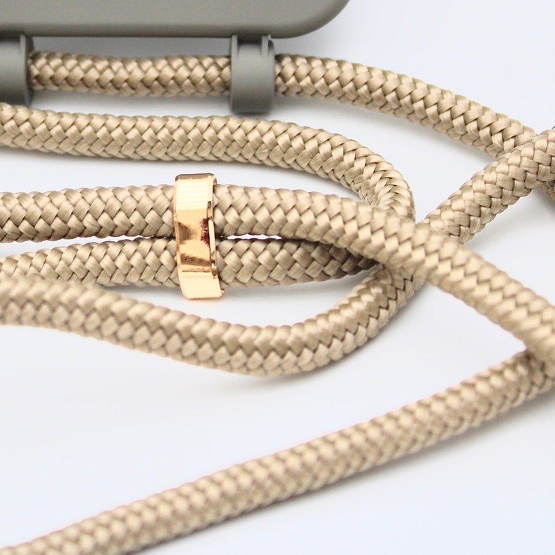 May include: Close-up of a beige woven phone strap with a gold-colored metal ring. The strap is attached to a gray phone case. The woven cord is thick and durable, and the gold ring adds a touch of elegance. The background is white.