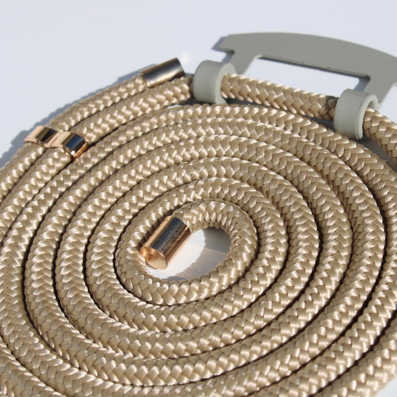 May include: A coiled, beige rope with gold-colored metal accents. The rope is thick and braided, with a gray plastic piece at the top. The rope is designed to be worn around the neck.