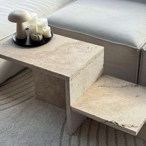 Könnte beinhalten: Ein zweistufiger, beigefarbener Beistelltisch aus Travertin mit rechteckiger Oberseite und Stufen-Design. Ein schwarzes Tablett oben hält weiße Kerzen, einen kleinen Kürbis und eine pilzförmige Dekoration. Der Tisch steht auf einem gemusterten Teppich.
