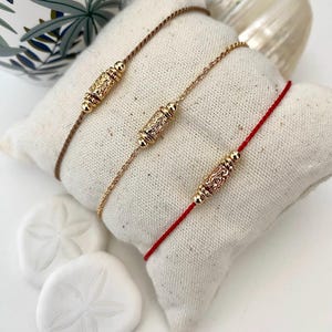 Puede incluir: Tres pulseras delicadas con cuentas cilíndricas doradas ornamentadas. Una pulsera tiene un cordón marrón, otra una cadena dorada y la tercera un cordón rojo. Las pulseras descansan sobre una almohada blanca, con dólares de arena blancos en primer plano.