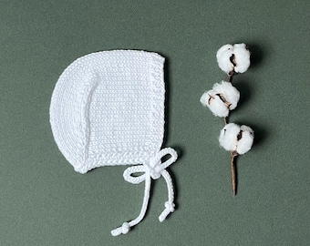 May include: A white knitted baby bonnet with a tie closure, next to a sprig of cotton bolls. The bonnet is made of a soft, textured yarn and has a rounded shape. The cotton bolls are fluffy and white, with a brown stem. The background is a muted green.
