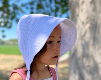 Peut inclure: Un jeune enfant portant un chapeau de soleil blanc à large bord et à liens. Le chapeau est fait d'un tissu doux et texturé. L'enfant porte une robe rose sans manches.