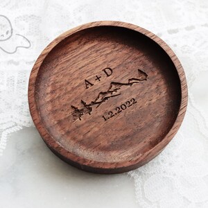 May include: A wooden ring dish with a personalised inscription that reads "A+D 1.2.2022" and features a mountain range and pine trees.