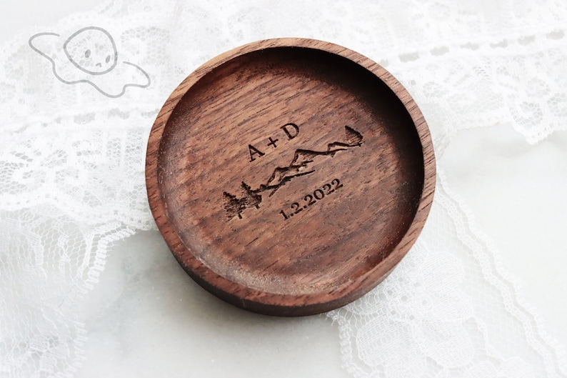 May include: A wooden ring dish with a personalised inscription that reads "A+D 1.2.2022" and features a mountain range and pine trees.