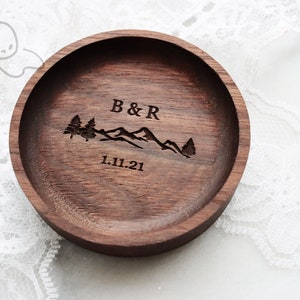 May include: A dark brown wooden ring dish with a mountain range and two trees engraved on the top. The text "B&R 1.11.21" is also engraved on the dish.