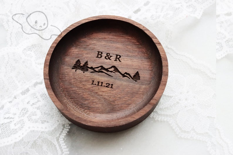 May include: A dark brown wooden ring dish with a mountain range and two trees engraved on the top. The text "B&R 1.11.21" is also engraved on the dish.