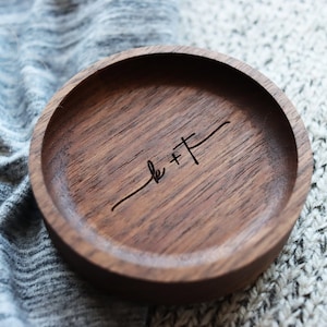 May include: A small, round wooden ring dish with a dark brown finish. The dish has a simple design and is engraved with the text "k + f".