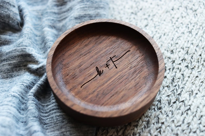 May include: A small, round wooden ring dish with a dark brown finish. The dish has a simple design and is engraved with the text "k + f".