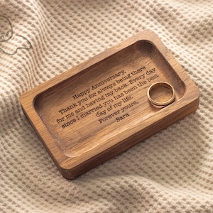 May include: A wooden ring dish with a personalized message engraved on the surface. The message reads "Happy Anniversary. Thank you for always being there for me and having my back. Every day since I married you has been the best day of my life. Forever yours, Sara". A gold ring is resting on the dish.