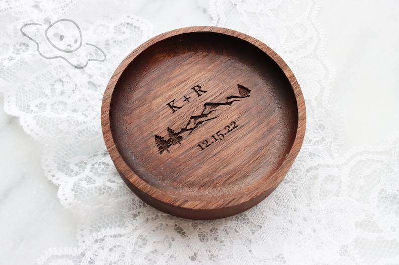 May include: A round wooden ring dish with a personalised inscription. The inscription reads "K+R" with a mountain range and trees, and the date "12.15.22".