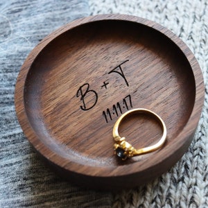 May include: A wooden ring dish with a dark brown finish. The dish is engraved with the text "B+T 11.11.17" and a gold ring with a black stone is sitting inside.