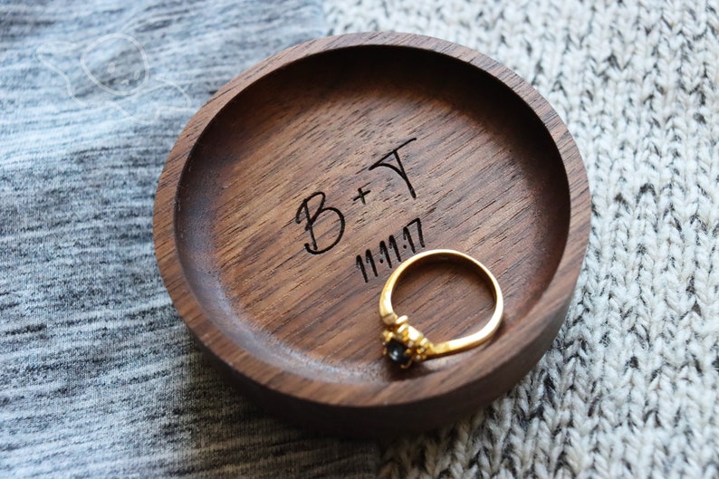 May include: A wooden ring dish with a dark brown finish. The dish is engraved with the text "B+T 11.11.17" and a gold ring with a black stone is sitting inside.