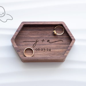 May include: A hexagonal wooden ring dish with a dark wood finish. The dish is engraved with the text "J + A" and "06.23.24". Two gold rings are placed inside the dish.