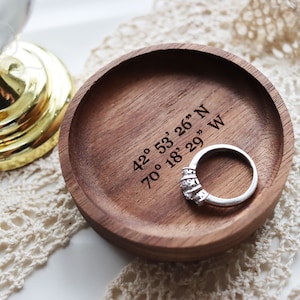 May include: A wooden ring dish with a ring inside. The dish is engraved with the coordinates "42° 53' 26" N 70° 18' 29" W".