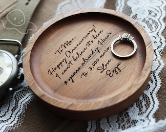 May include: A wooden ring dish with a personalized message: "To Max. Happy Anniversary! I can't believe it's been 3 years already. Here's to 3,000 more. Love, Ezgu". A silver ring with three small diamonds is resting on the dish.